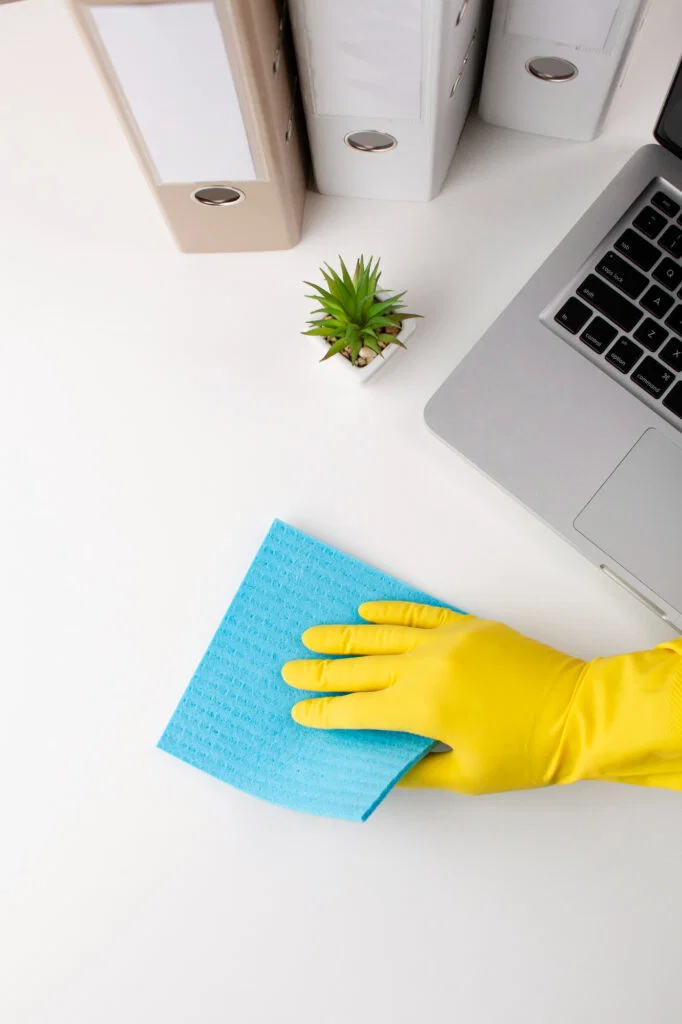 Hand in yellow glove cleaning desk near laptop, plant, and folders with a blue cloth—ideal for thorough office cleaning.
