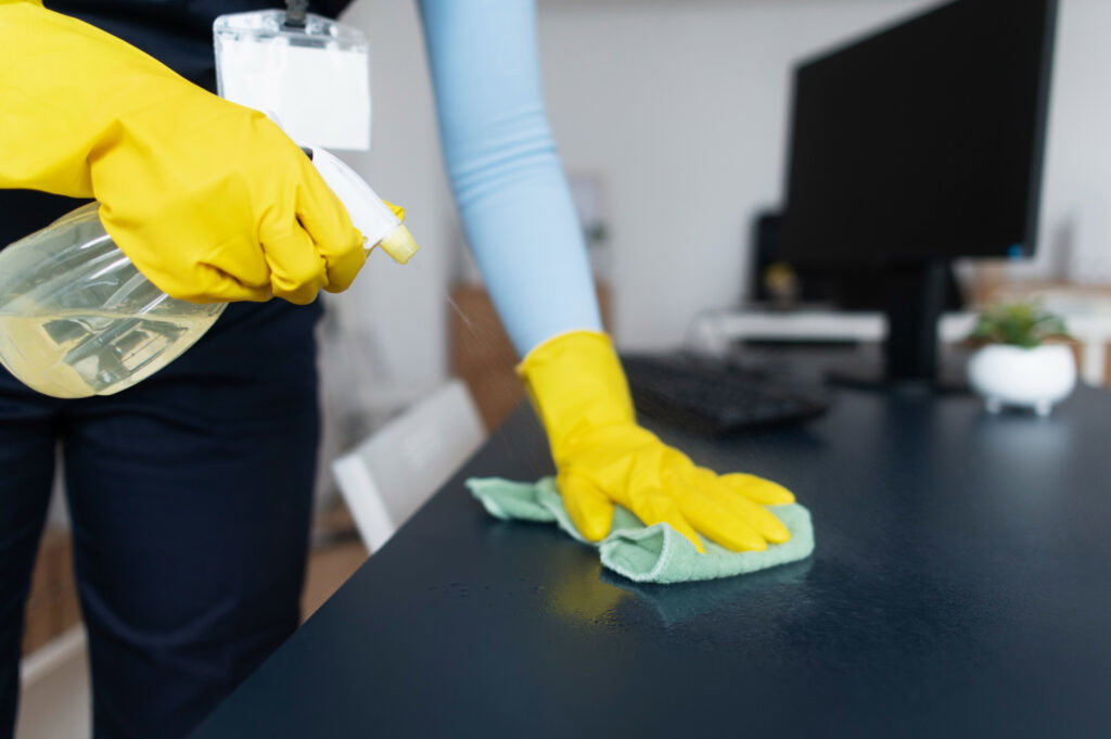 Person in yellow gloves cleaning a desk with a spray bottle and cloth in an office setting, showcasing the expertise of a professional deep cleaning contractor.