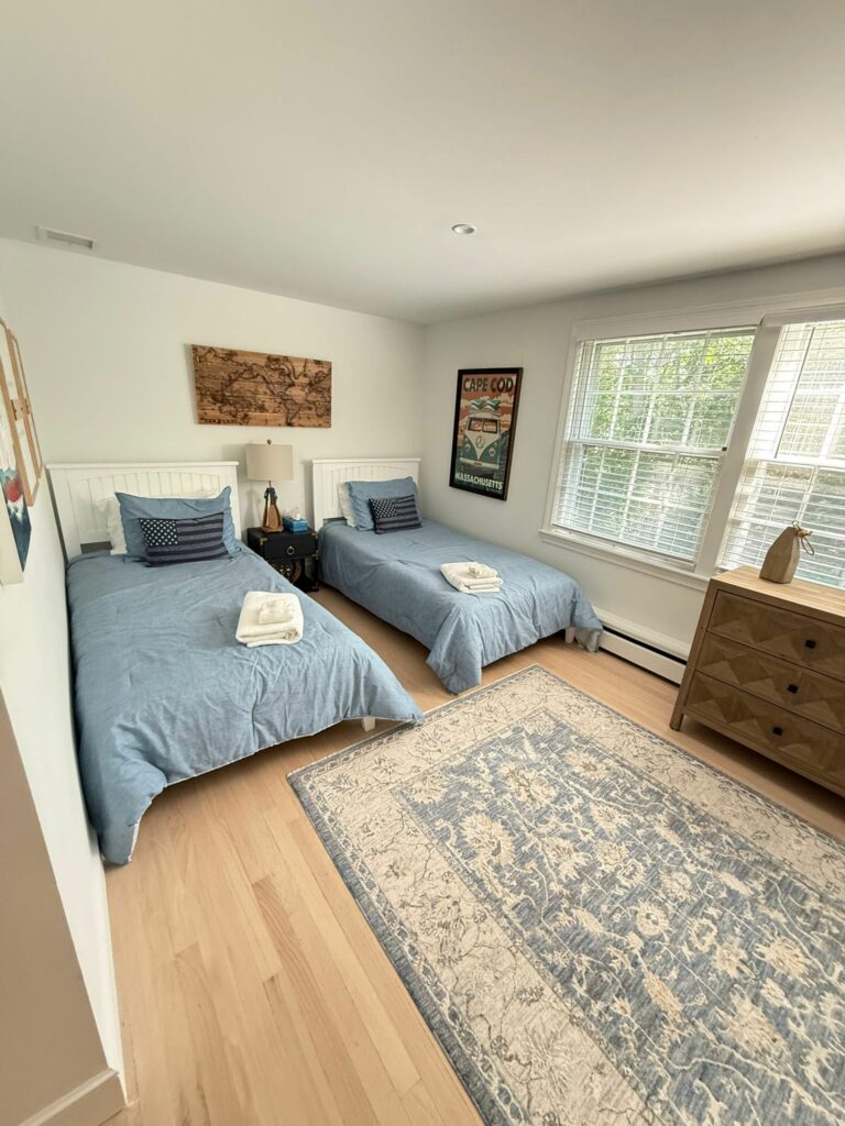 Two twin beds with blue bedding in a bright bedroom featuring large windows, art, and a patterned rug—kept spotless by an Airbnb cleaner for your comfort.