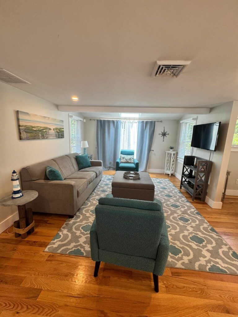 Living room with teal and gray furniture, patterned rug, wall art, TV, hardwood floor, lighthouse decor, and a fresh feel thanks to recent professional home cleaning.