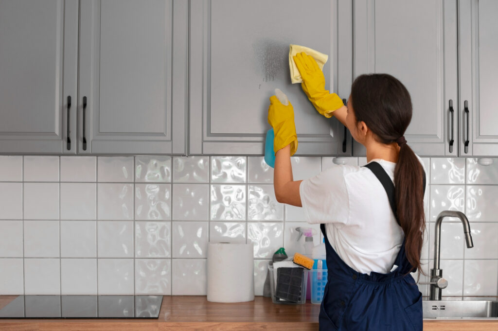 Woman wearing yellow gloves cleaning a gray kitchen cabinet with a sponge and spray bottle, just like a professional Weekly Maid keeping your home spotless.