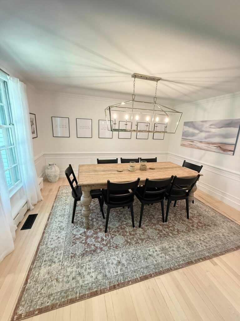 A dining room with a wooden table, black chairs, wall art, a rug, and a modern chandelier—kept spotless with the help of a Monthly Maid service.